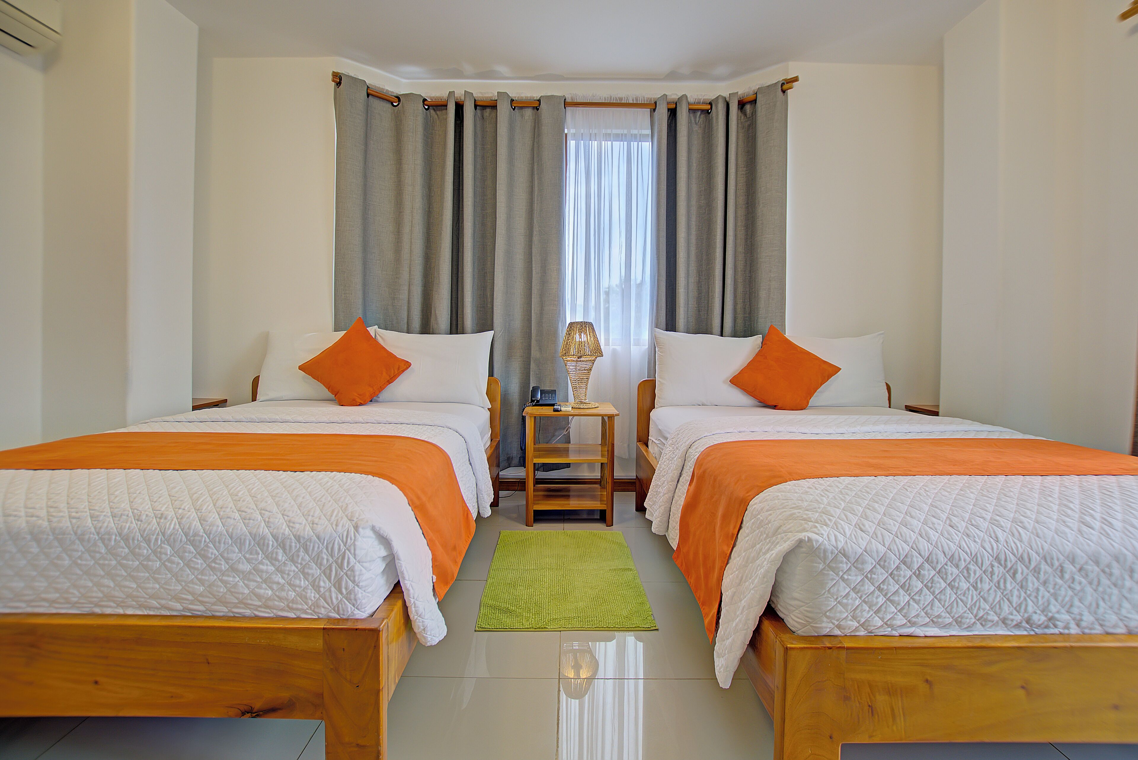Une chambre double avec deux lits, chacun agrémenté de draps blancs et orange, un tapis vert et une petite table avec une lampe placée entre les lits.