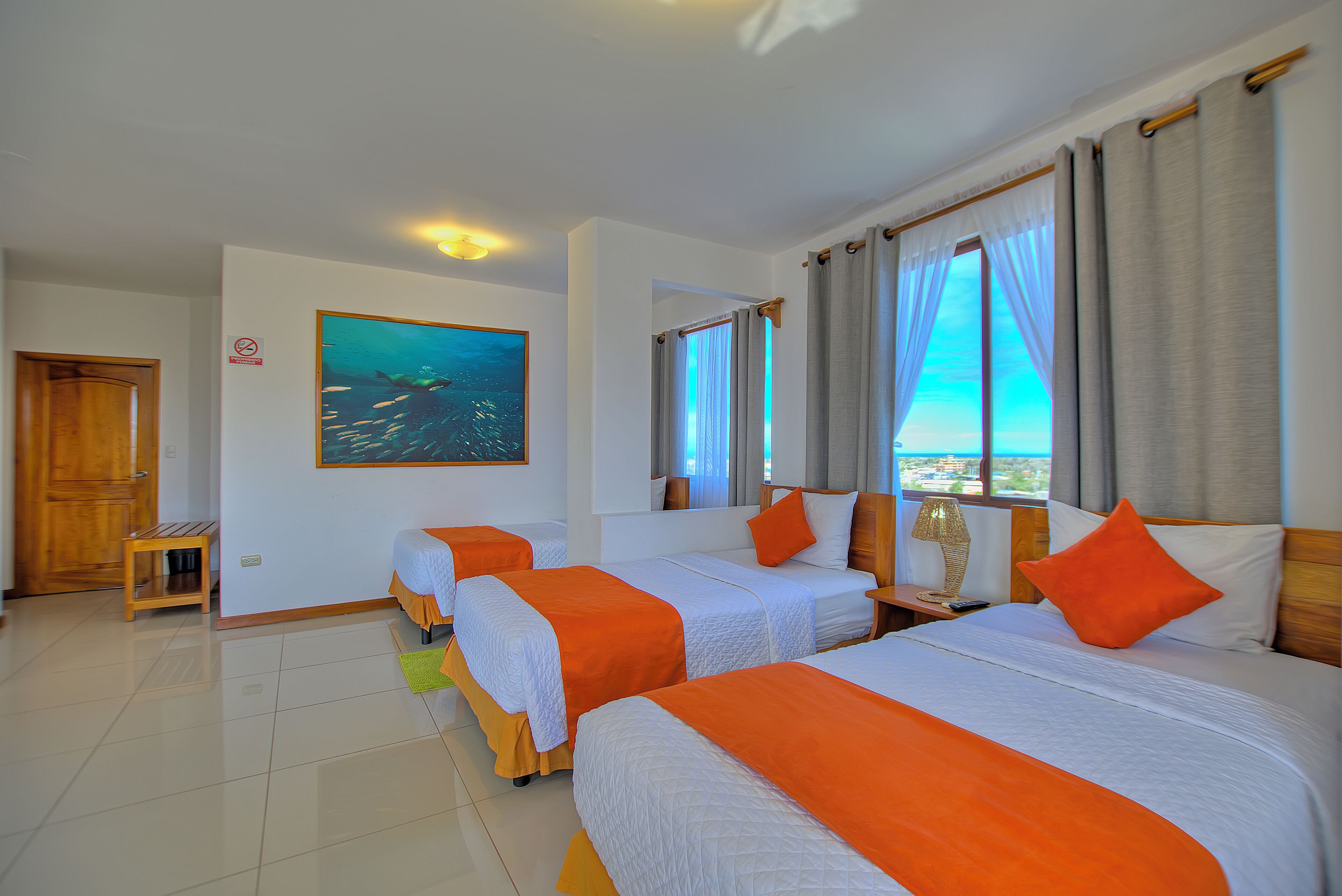 A hotel room with three beds, a painting of fish on the wall, and a view of the sea through the window.