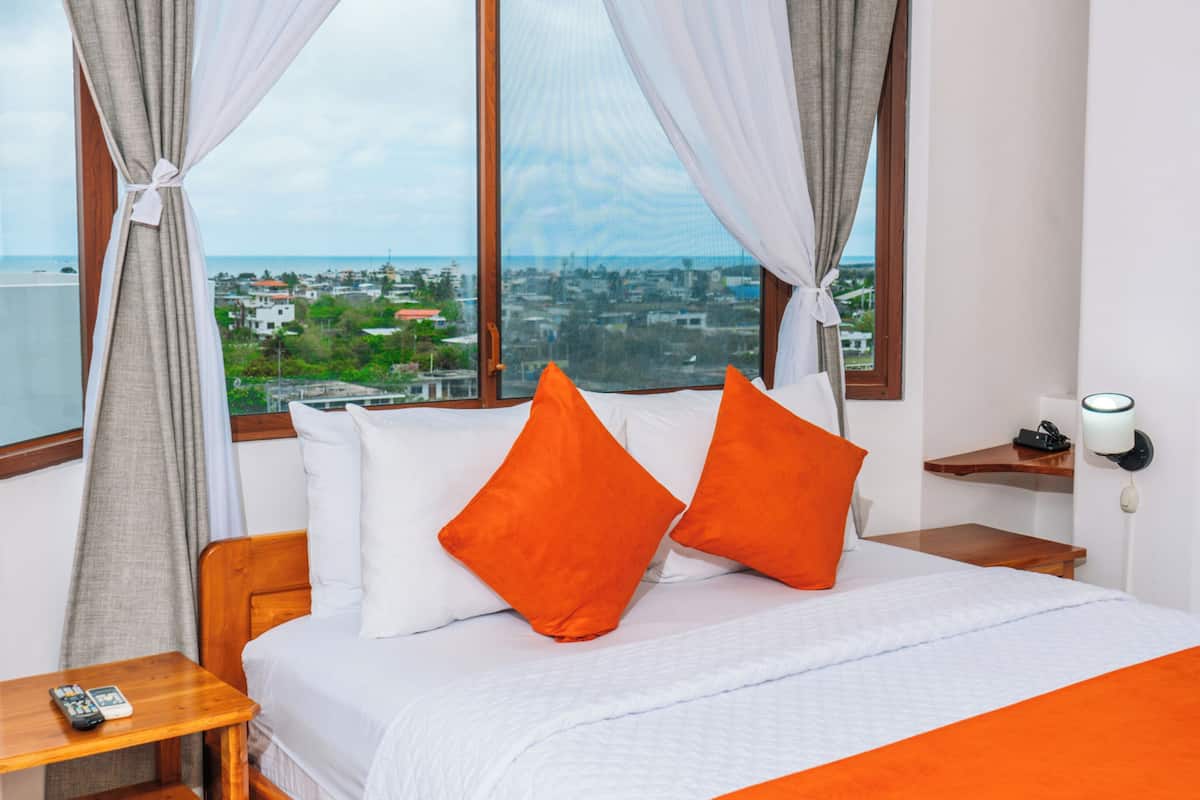 Une chambre d’hôtel avec un lit, deux oreillers orange, une table de chevet en bois, une télécommande et une vue sur le paysage urbain à travers de grandes fenêtres.
