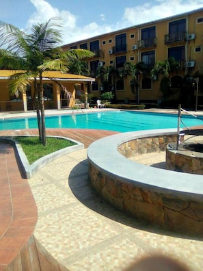 Una piscina con un hogar de piedra, rodeada por una terraza con baldosas y palmeras.