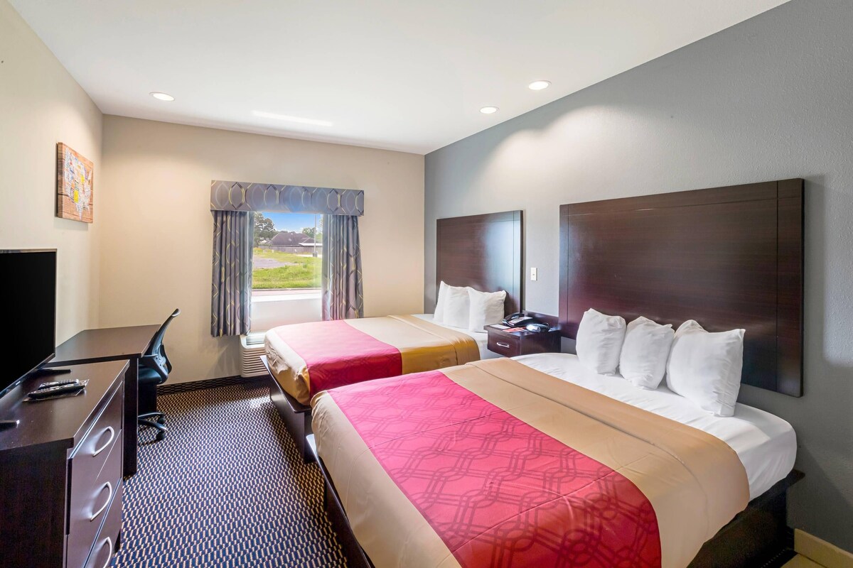 A hotel room with two beds, a desk, a chair, a TV, and a view of the outdoors.