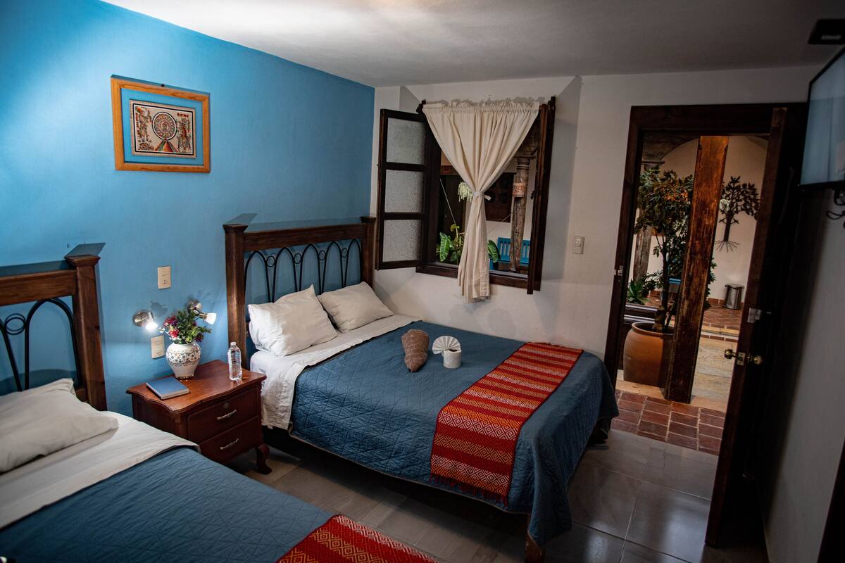 Quarto com duas camas, uma mesa de cabeceira com um laptop e um vaso, uma janela com cortina e uma porta que dá para um corredor.