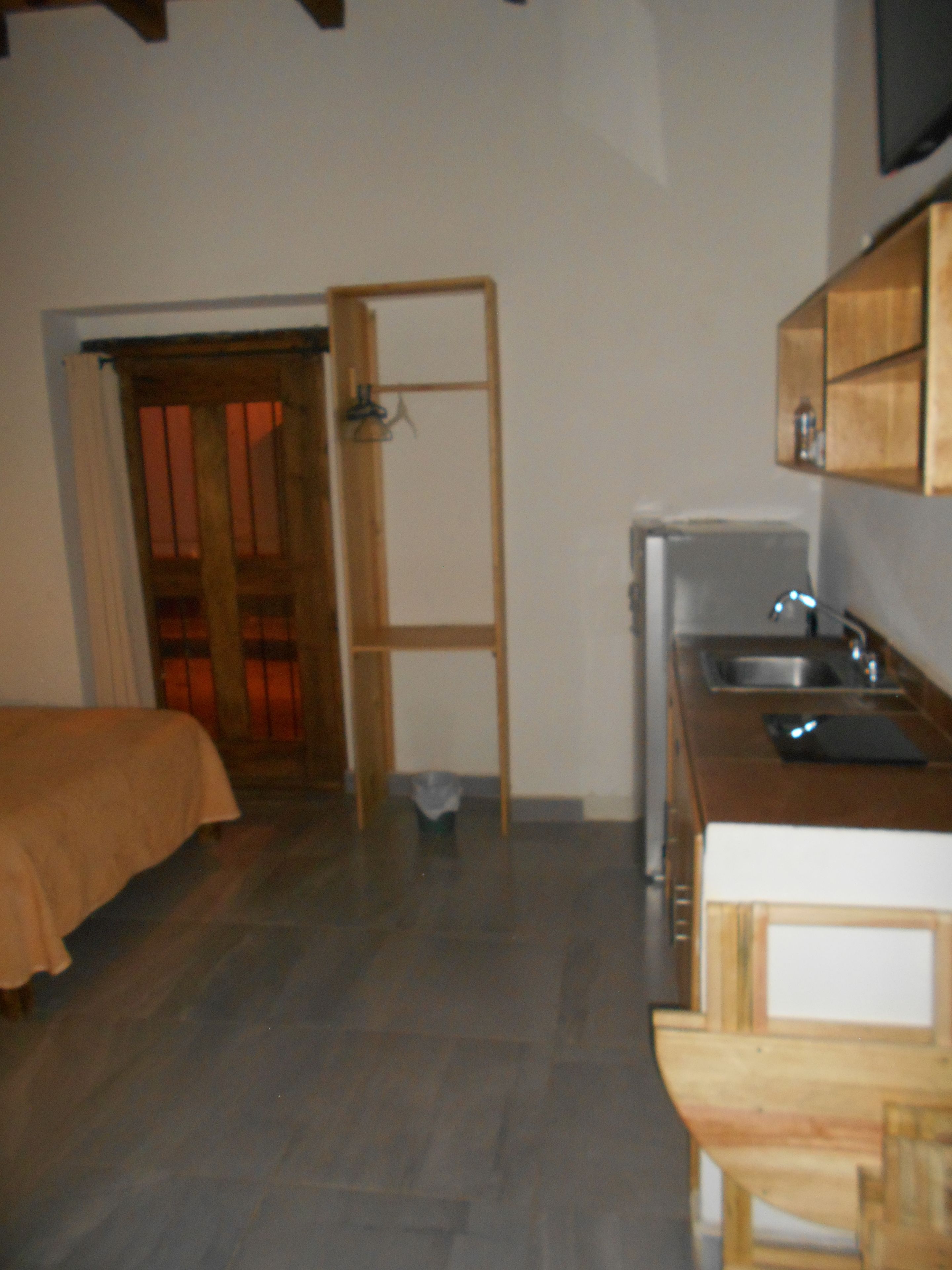 A small, compact kitchen with a refrigerator, sink, and wooden cabinets. There is a bed with a brown cover in the adjacent room.