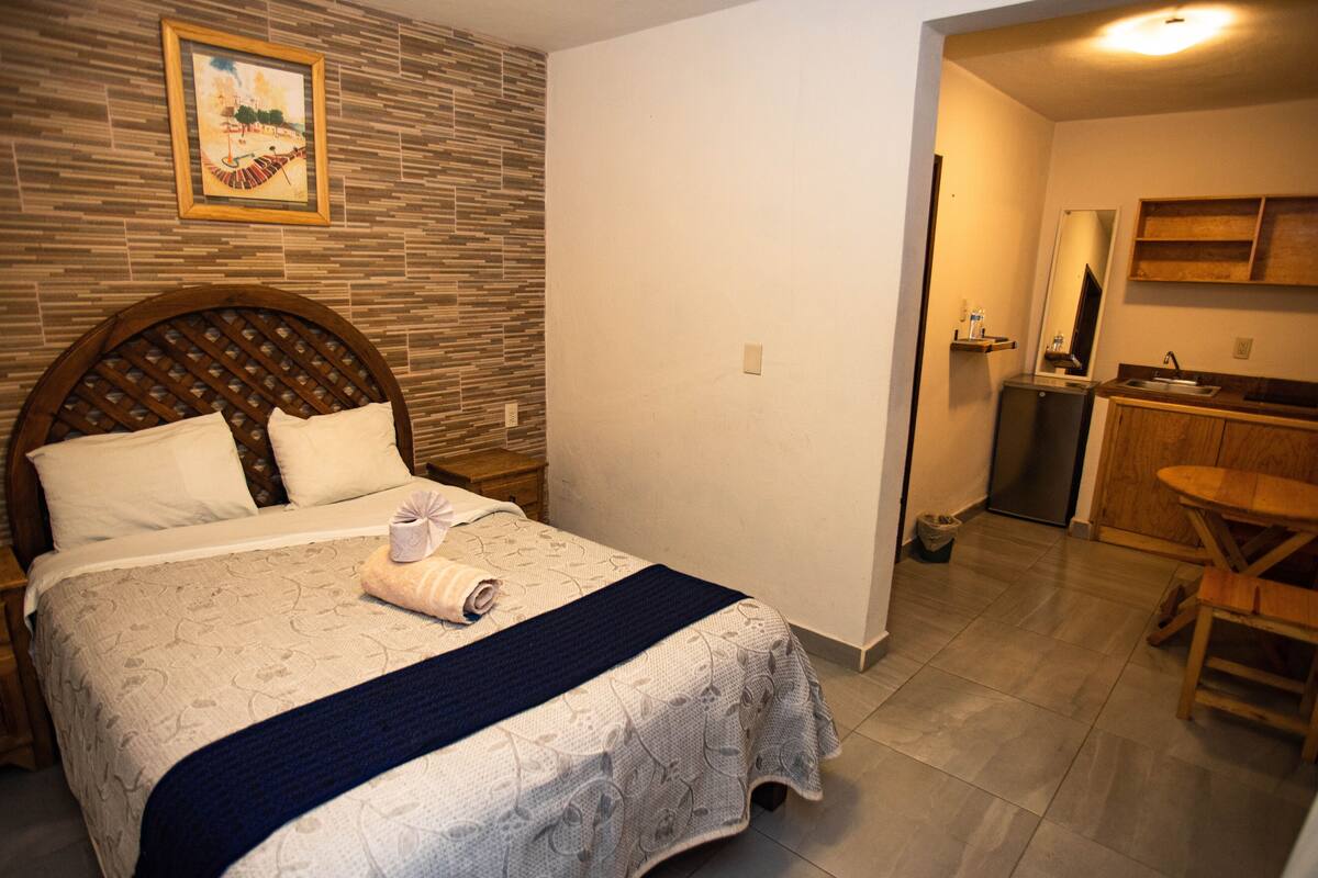 Quarto bem arrumado com uma cama grande, cabeceira de madeira, mesa de cabeceira pequena e um criado-mudo. É visível um banheiro com pia, espelho e um pequeno nicho.