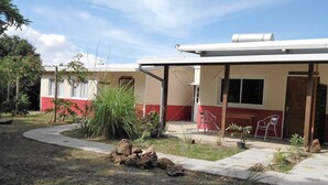 Un edificio con fachada roja y blanca, un porche cubierto con sillas y un jardín con rocas y plantas.