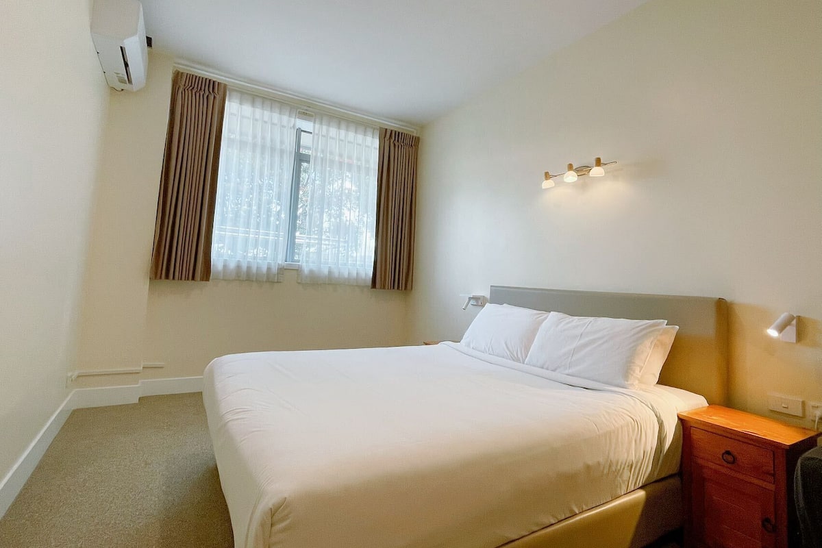 A hotel room with a bed, a nightstand, a lamp, and a window with curtains.