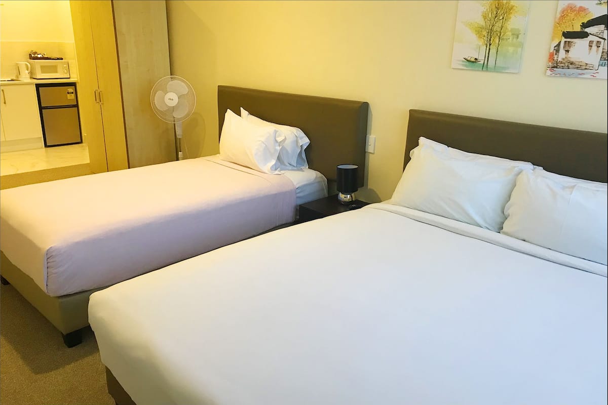 Two single beds with white linens, a bedside table with a lamp, a wall-mounted fan, and a microwave on the left.