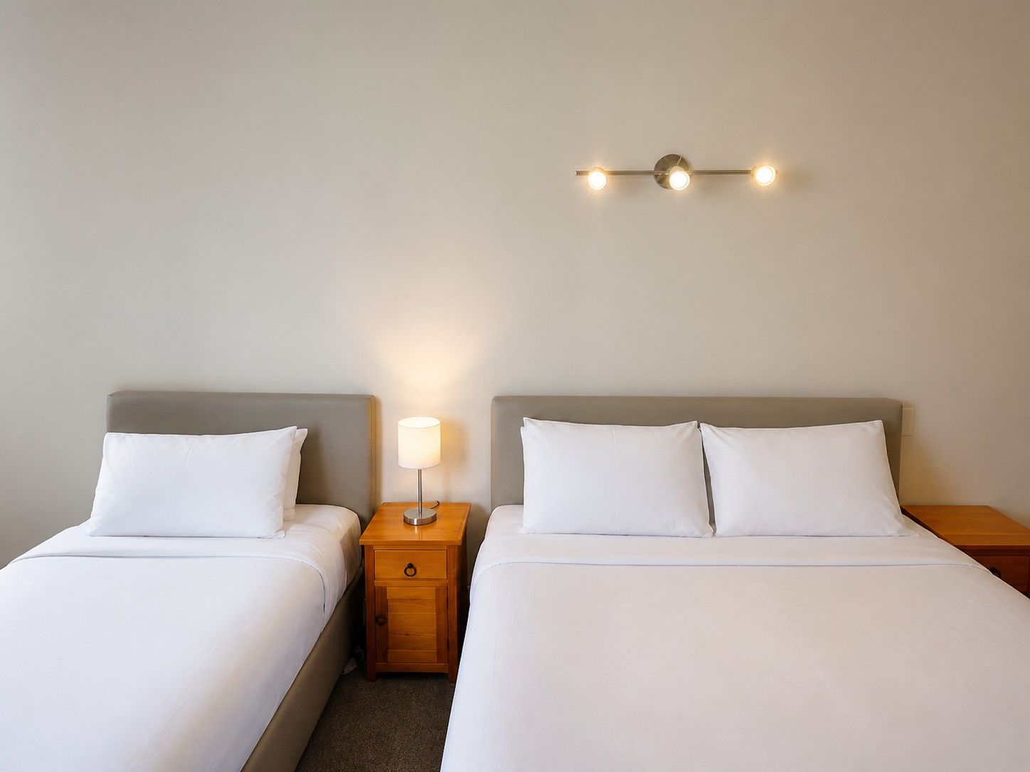 A hotel room with two beds, a nightstand with a lamp, and a wall-mounted light fixture.