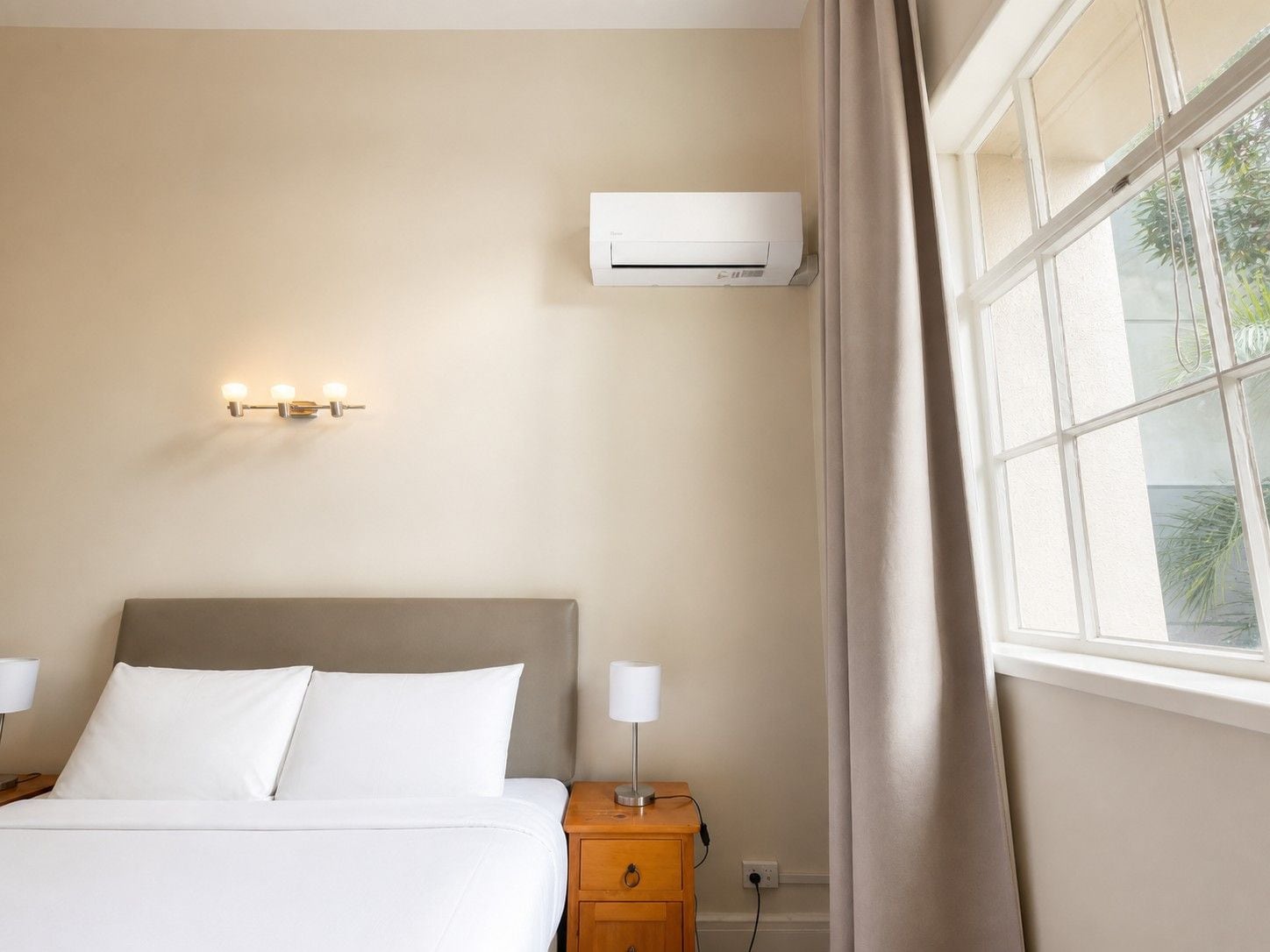 Habitación de hotel con cama, mesita de noche, lámpara, equipo de aire acondicionado y ventana con cortinas.