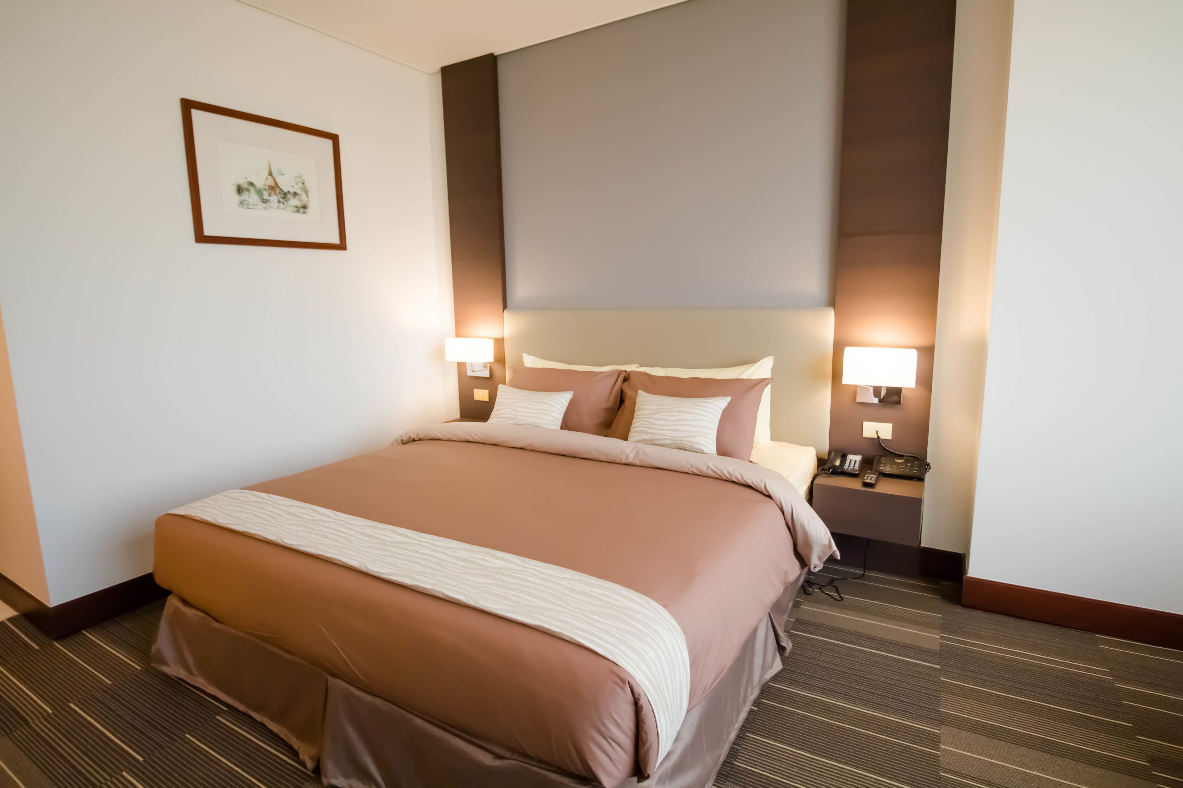 A hotel room with a large bed, two bedside lamps, a framed picture on the wall, and a carpeted floor.