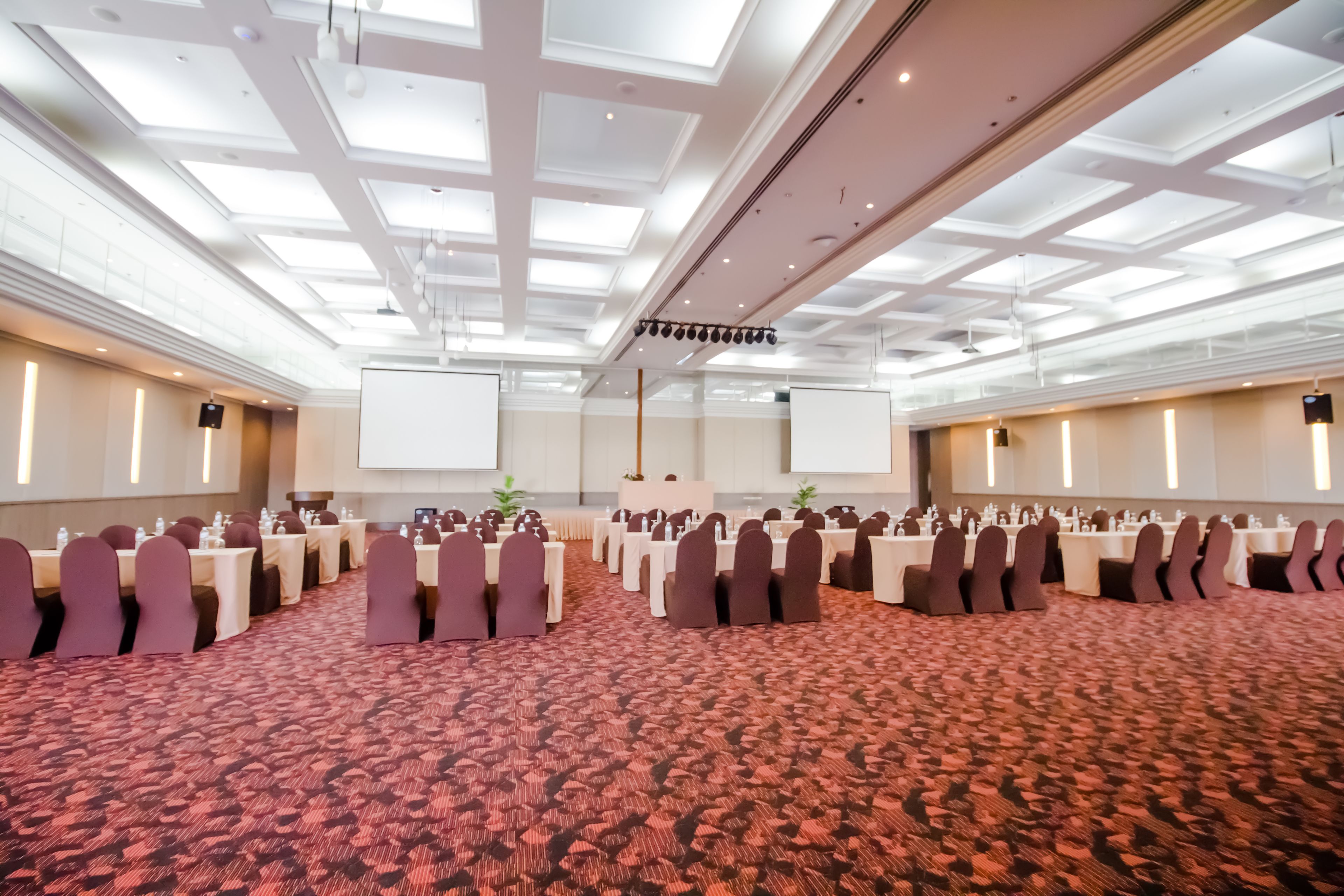 A large conference room with rows of tables and chairs, a projector screen, and a carpeted floor.