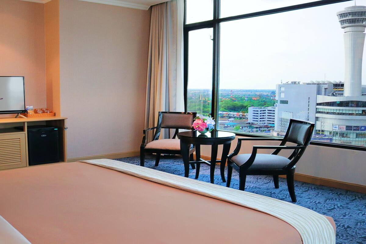 A hotel room with a bed, a desk with a television displaying the Fortune Hotel Group logo, a small table with chairs, and a large window offering a city view.