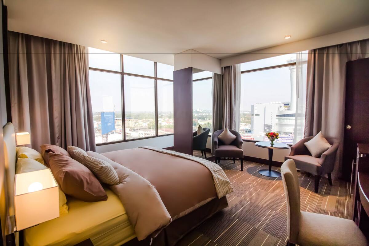 A hotel room with a large bed, two armchairs, a small table, and a view of the city.