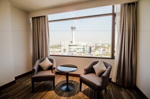Une chambre d’hôtel avec deux fauteuils et une petite table, ainsi qu’une grande fenêtre donnant sur une tour et le paysage urbain.