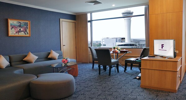Habitación de hotel con sofá, reposapiés y escritorio con televisor. Se ve una ventana con vista a una torre y un aeropuerto.