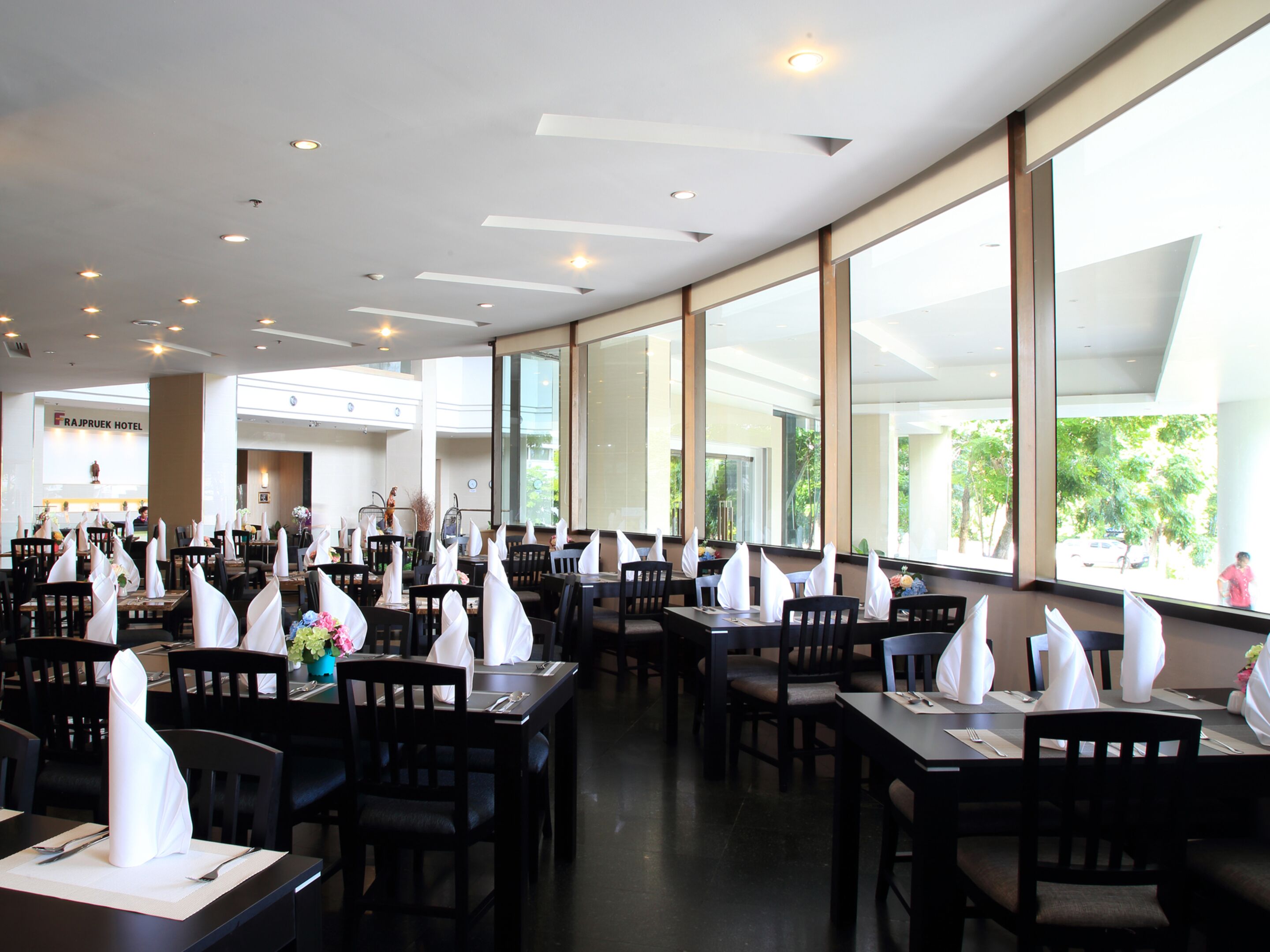 A spacious restaurant with high ceilings and large windows, featuring neatly arranged tables with white napkins and chairs.
