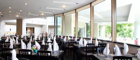 A spacious restaurant with high ceilings and large windows, featuring neatly arranged tables with white napkins and chairs.