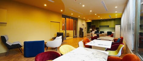 A modern dining area with round tables and colorful chairs, a kitchen in the background, and a bright, yellow-walled room.