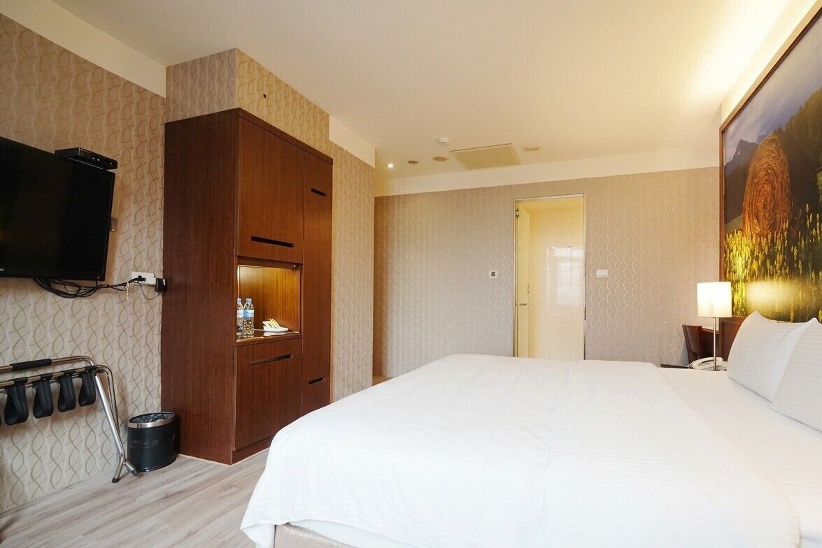 Quarto de hotel com uma cama grande, um guarda-roupa, uma televisão na parede, um quadro e uma porta que dá para outro cômodo.