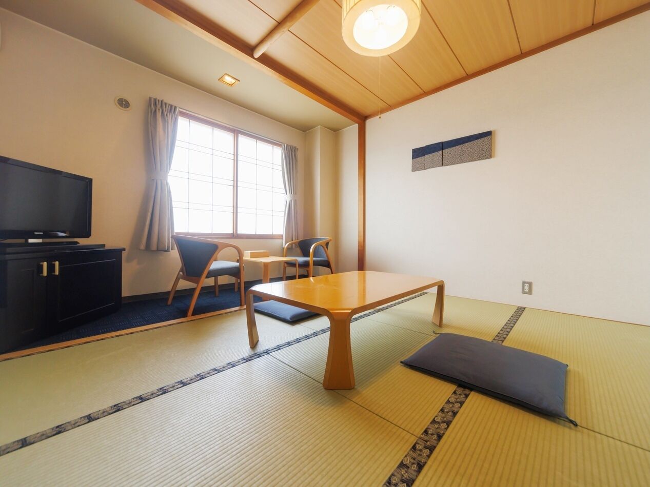 Una camera tradizionale giapponese con pavimento in tatami, un tavolino, due sedie, una televisione e una finestra con tende.