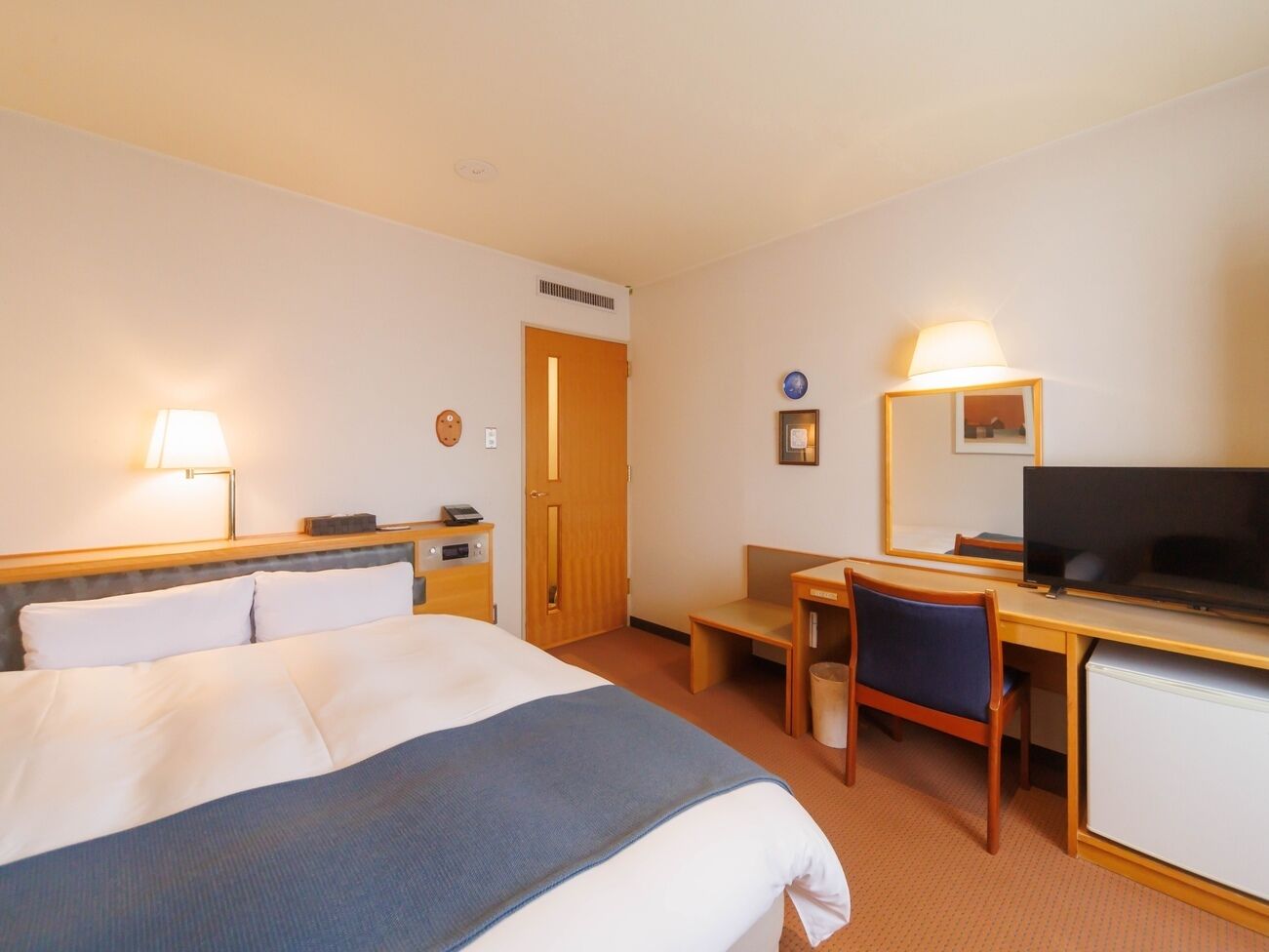 Una camera d'albergo con un letto, una scrivania, una sedia, una televisione e una porta.