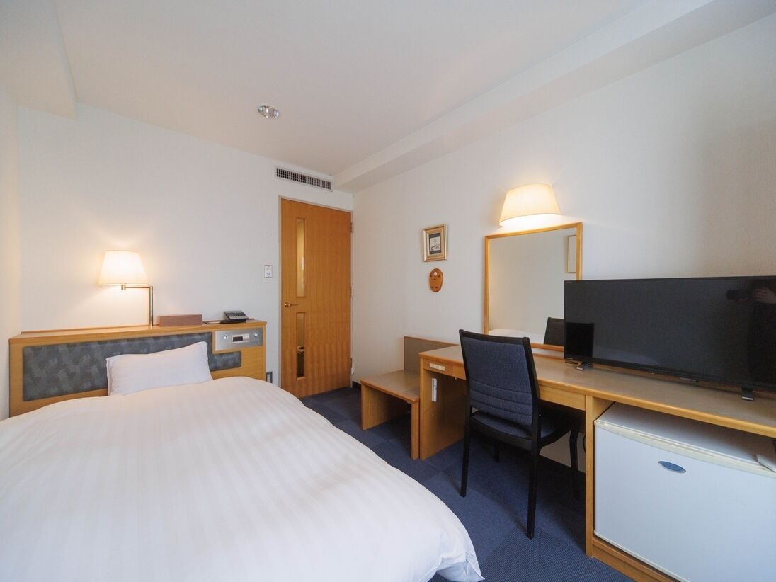Una camera d'albergo con un letto, una scrivania, una sedia, televisione e un piccolo frigorifero.
