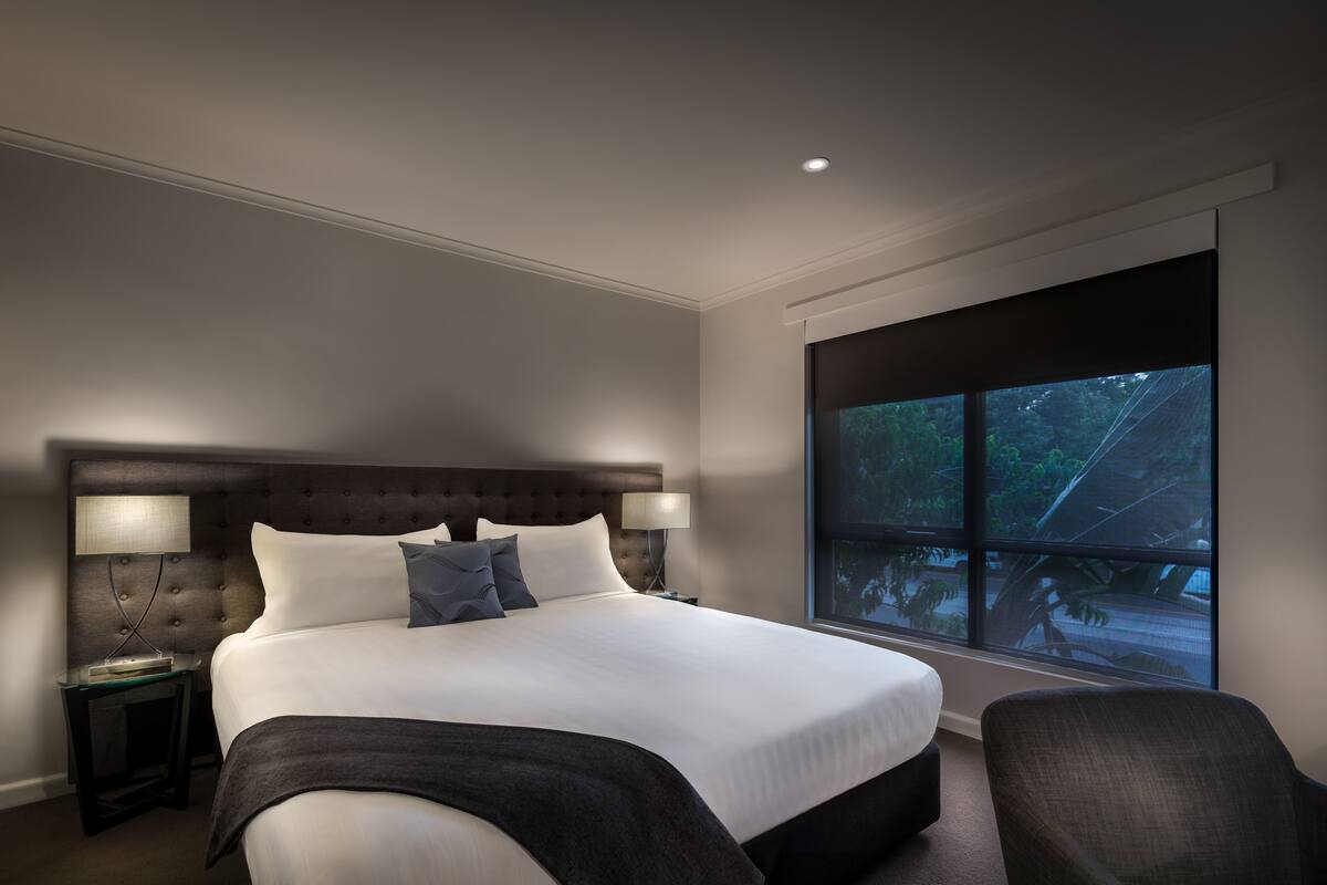 A large bed with a tufted headboard, two bedside tables with lamps, a chair, and a large window with a view of trees.