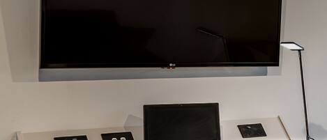 A modern room with a flat-screen TV mounted on the wall, a laptop on a white desk, and a desk lamp.