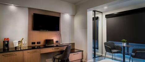 A modern hotel room with a flat-screen TV mounted on the wall, a desk with a chair, a small table with two chairs, and a large window offering a view of the city skyline.