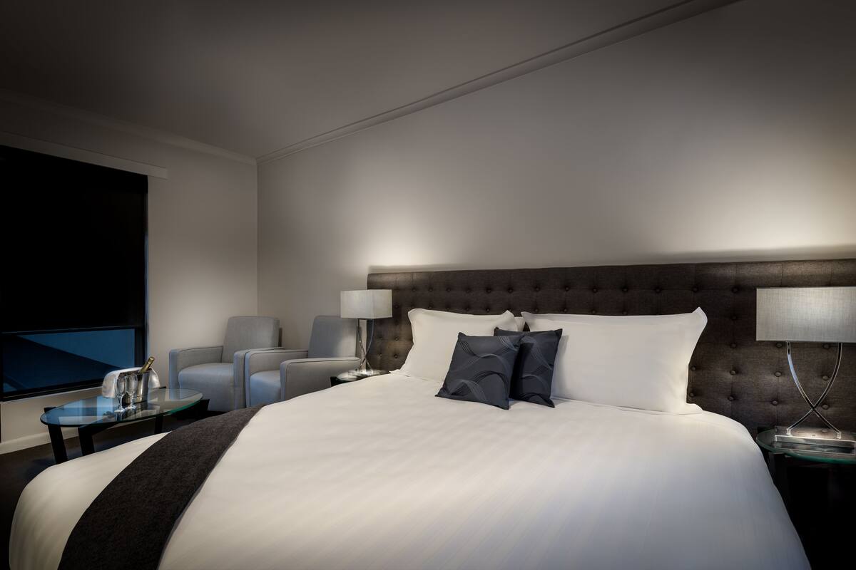 A modern hotel room with a large bed, a flat-screen TV, a seating area with two chairs, and a nightstand with a lamp.