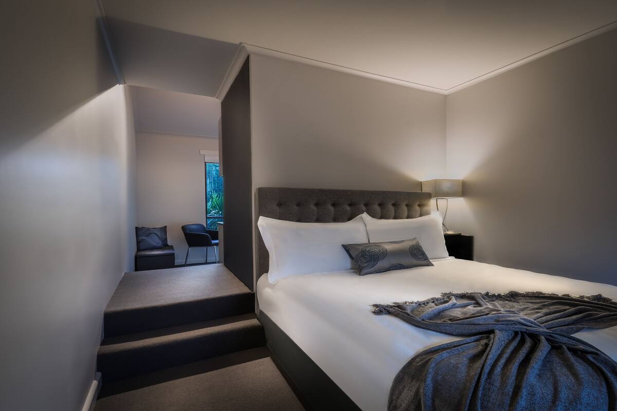 A modern hotel room with a large bed, a headboard, and a bedside table with a lamp. There is a staircase leading to an adjacent room.