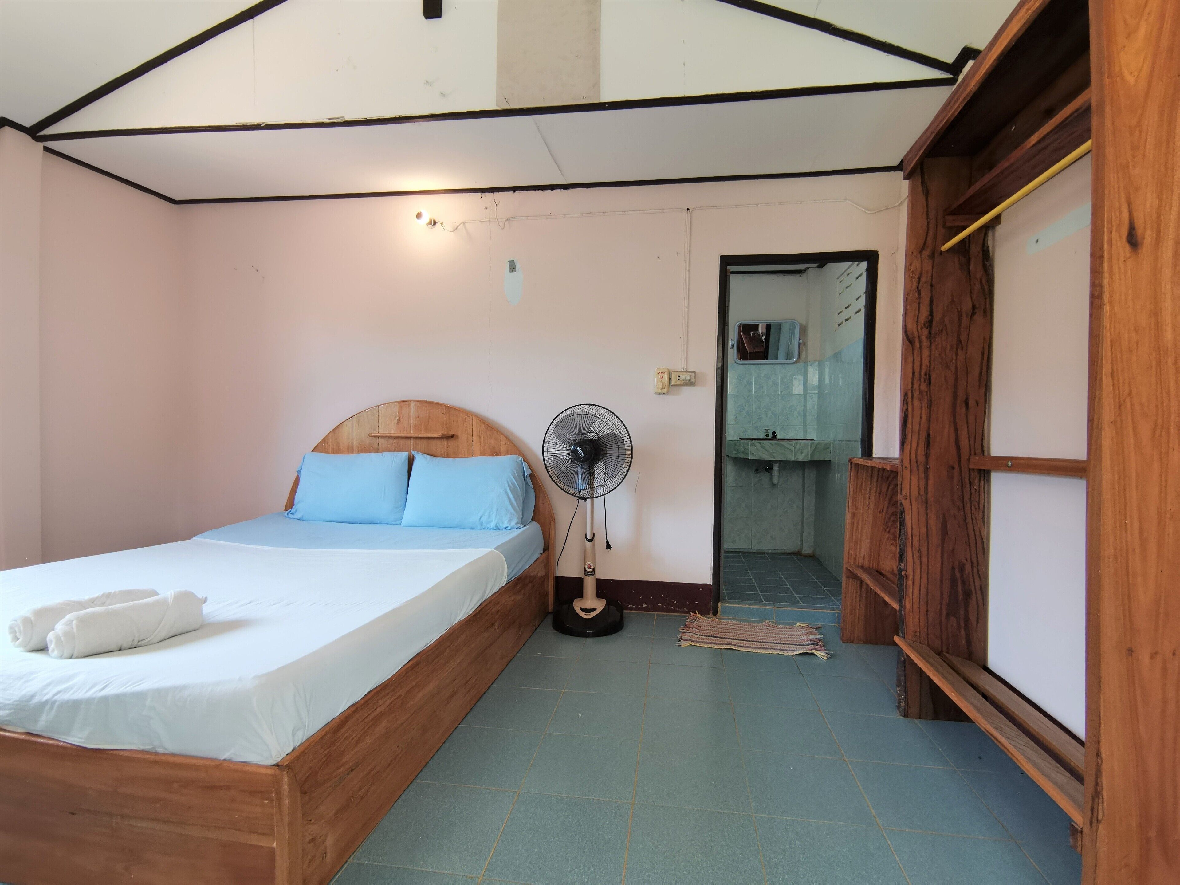 A bedroom with a wooden bed, a standing fan, and a bathroom visible through an open door.