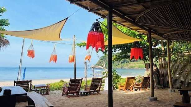 Un área de descanso en la playa con sillas y mesas de madera, protegida por una estructura con tela roja y naranja.