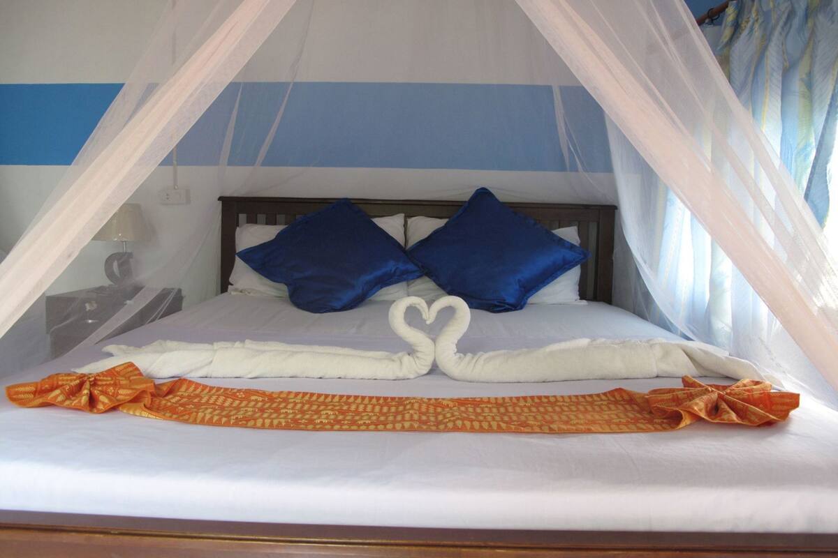 Een bed met hemel, twee kussens, een handdoek creatie in de vorm van een hart, en een bedekking met een patroon.