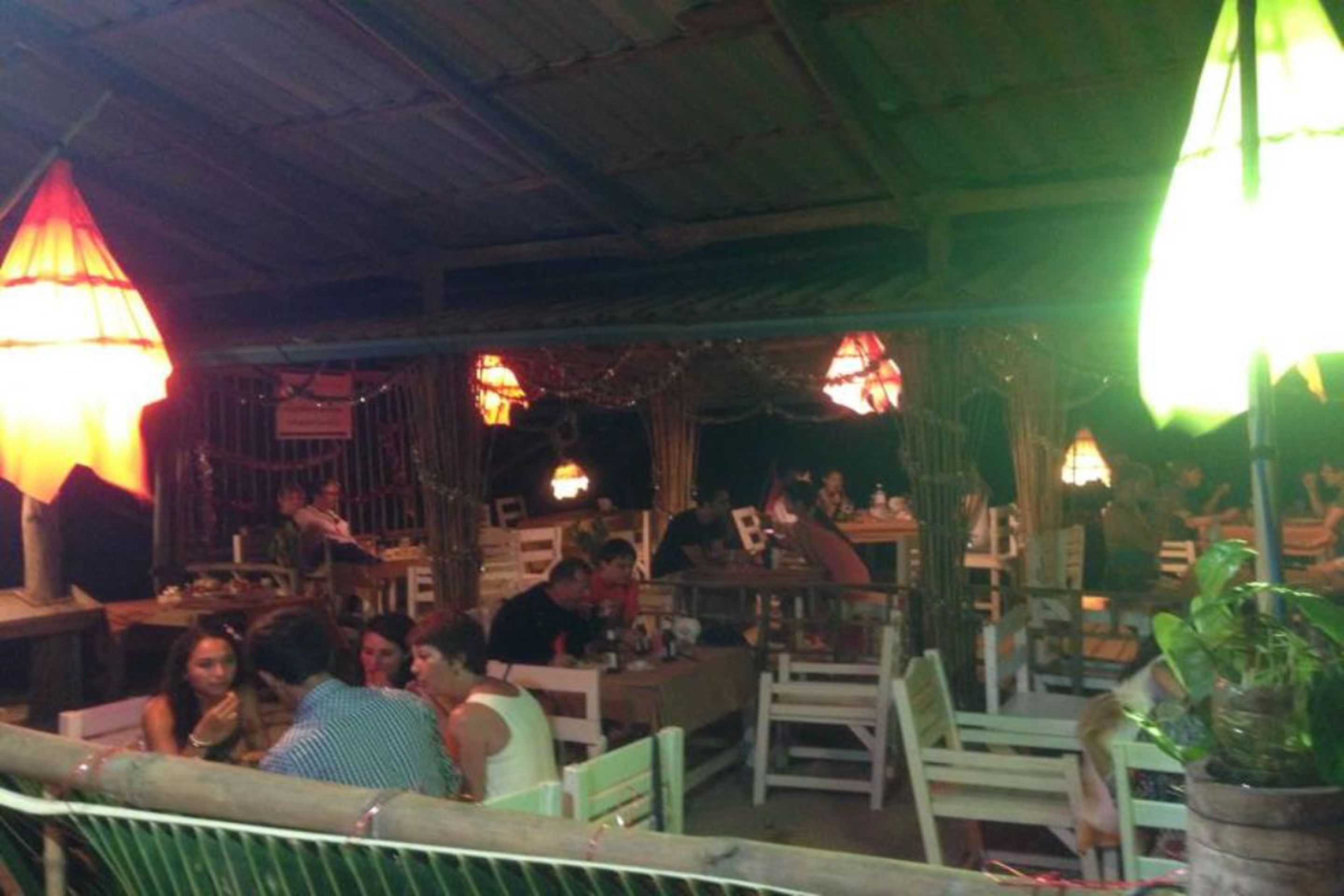 Un restaurante al aire libre con mesas y sillas de madera, lámparas colgantes y un techo rústico.
