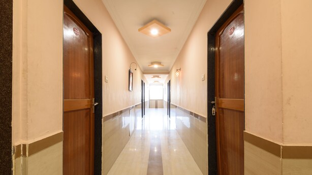 A corridor with wooden doors and a tiled floor, leading to a bright area at the end. - Hotel Sai Shraddha (Rahata)