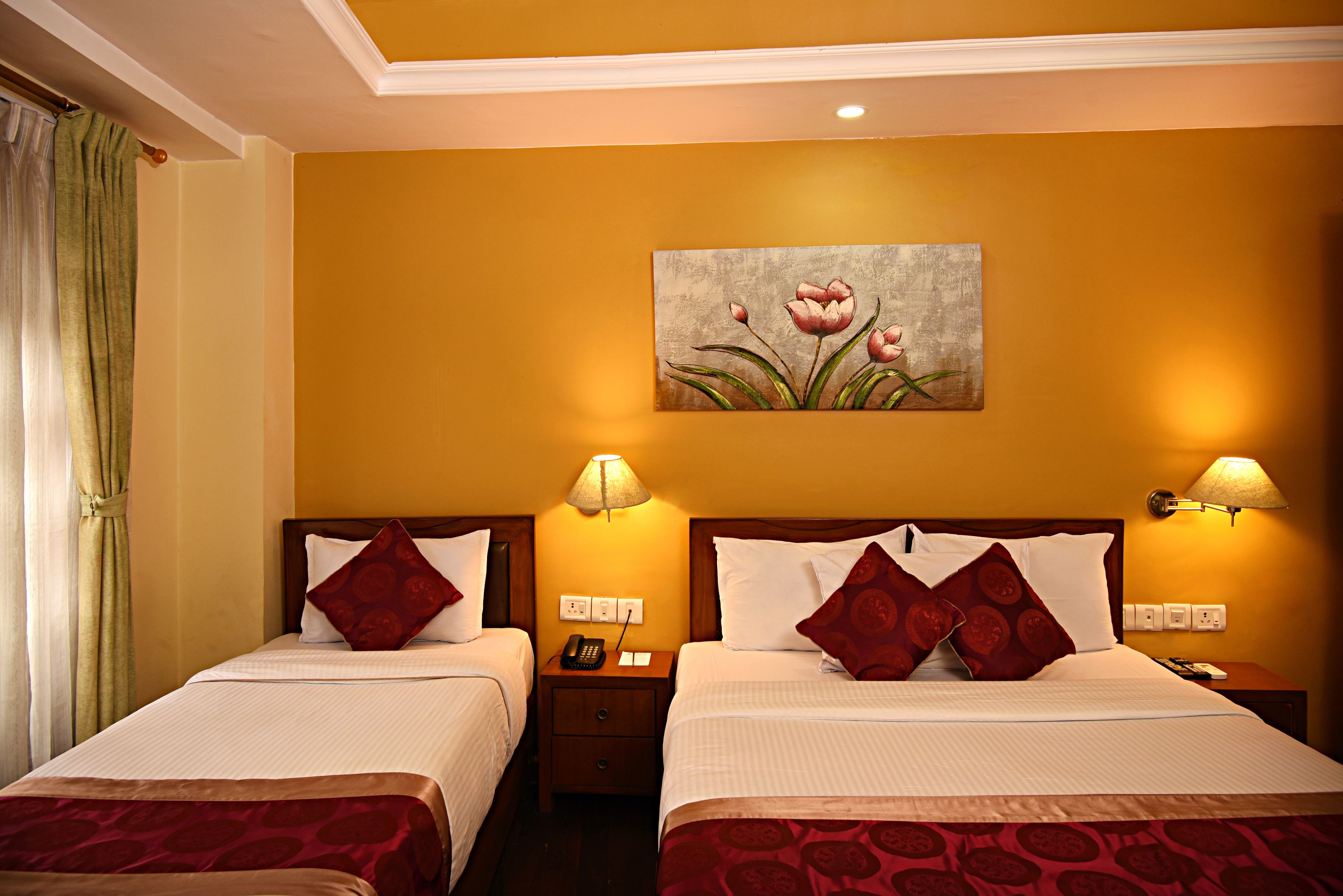 A hotel room with two beds, each with a red patterned bedspread, white linens, and matching red pillows. There is a painting of flowers on the wall, a nightstand with a lamp, and a telephone.