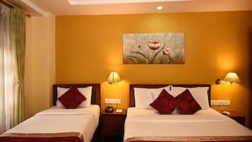 A hotel room with two beds, each with a red patterned bedspread, white linens, and matching red pillows. There is a painting of flowers on the wall, a nightstand with a lamp, and a telephone.