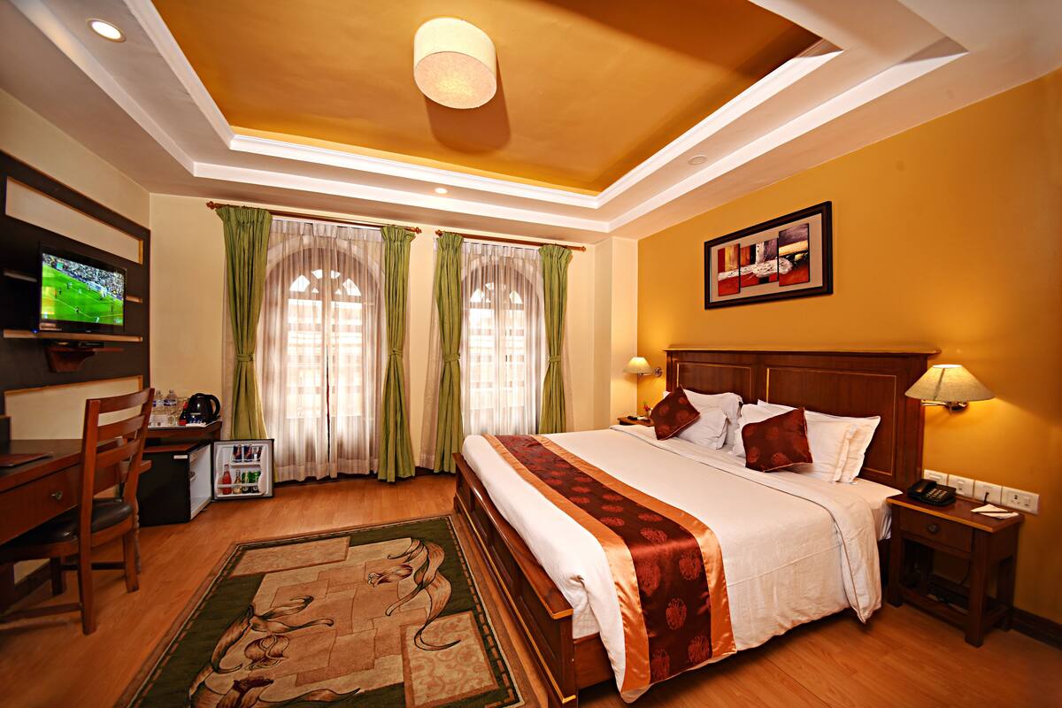 A hotel room with a large bed, a desk with a television, a small refrigerator, and a wooden nightstand with a lamp.
