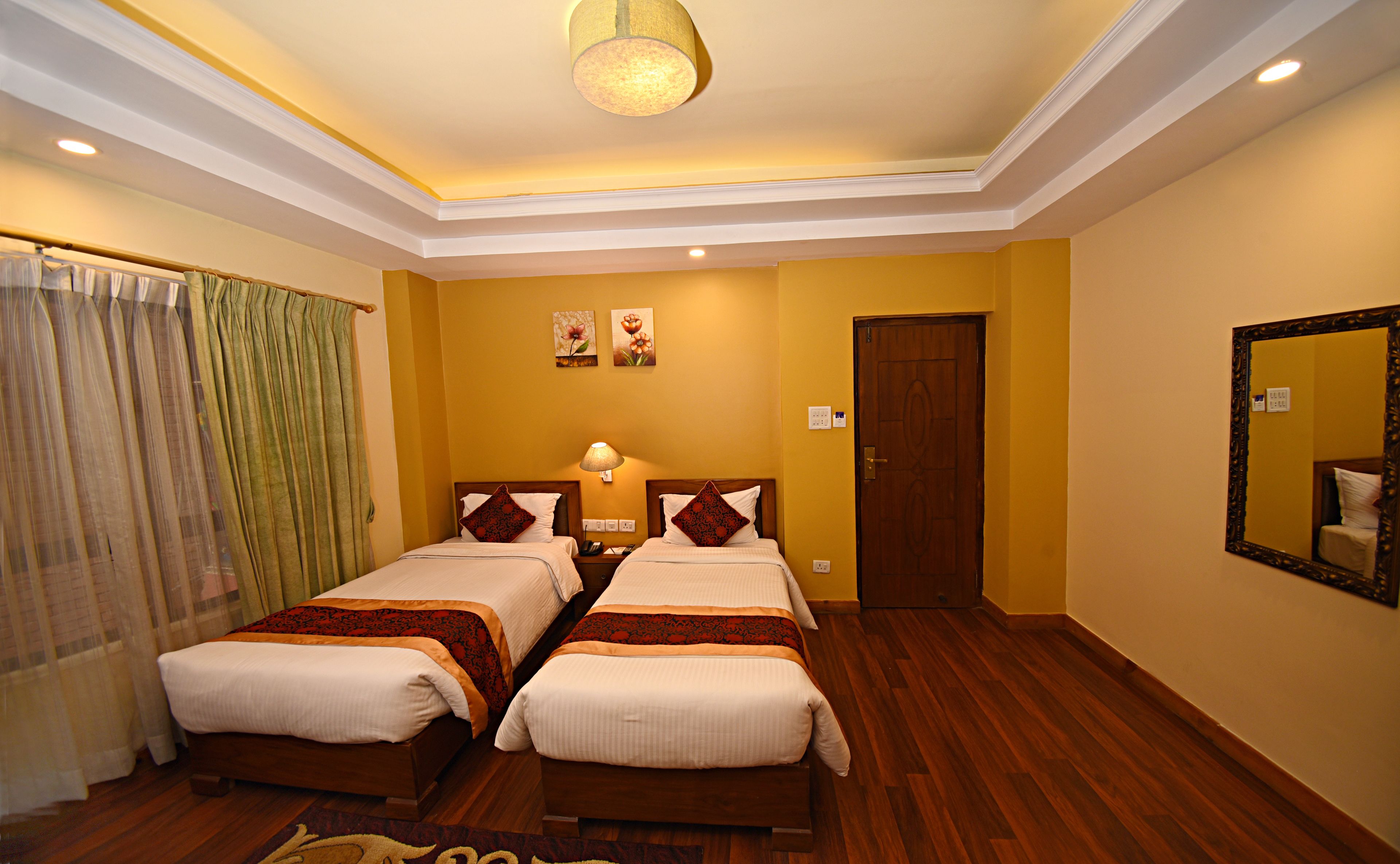 A hotel room with two beds, a wooden floor, a ceiling light, a door, and a mirror on the wall.