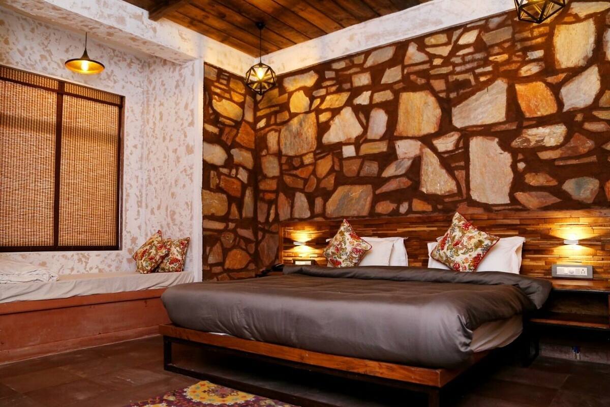 Un dormitorio con pared de piedra, cama con estructura de madera, ventana con persiana tejida y dos lámparas fijadas a la pared.