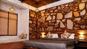 A bedroom with a stone wall, a bed with a wooden frame, a window with a woven blind, and two wall-mounted lights.