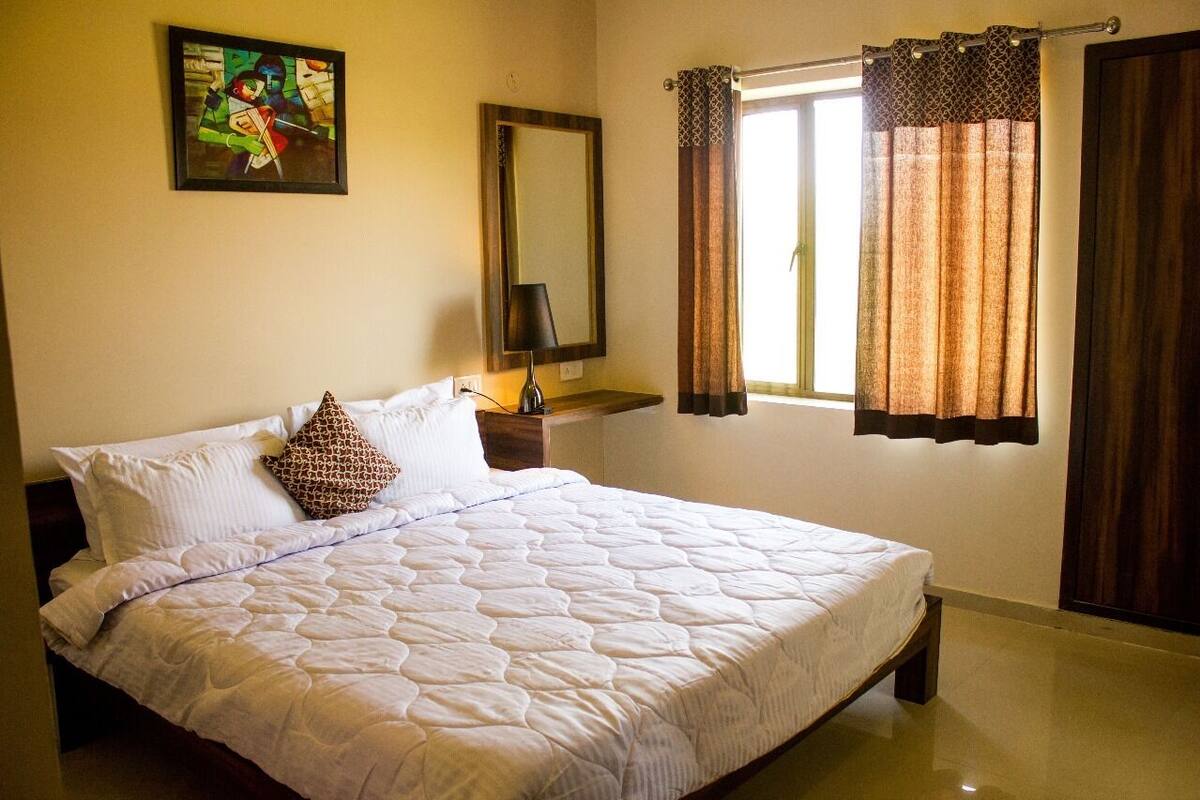 Una cama bien hecha con edredón blanco acolchado, una única almohada marrón estampada y una mesita de noche con lámpara. Hay una ventana con cortinas marrones y un cuadro en la pared sobre la cama.