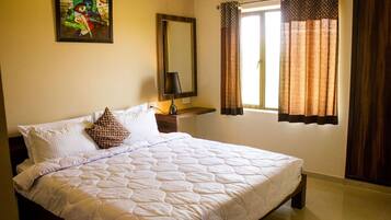A neatly made bed with a quilted white duvet, a single brown patterned pillow, and a bedside table with a lamp. There is a window with brown curtains and a painting on the wall above the bed.