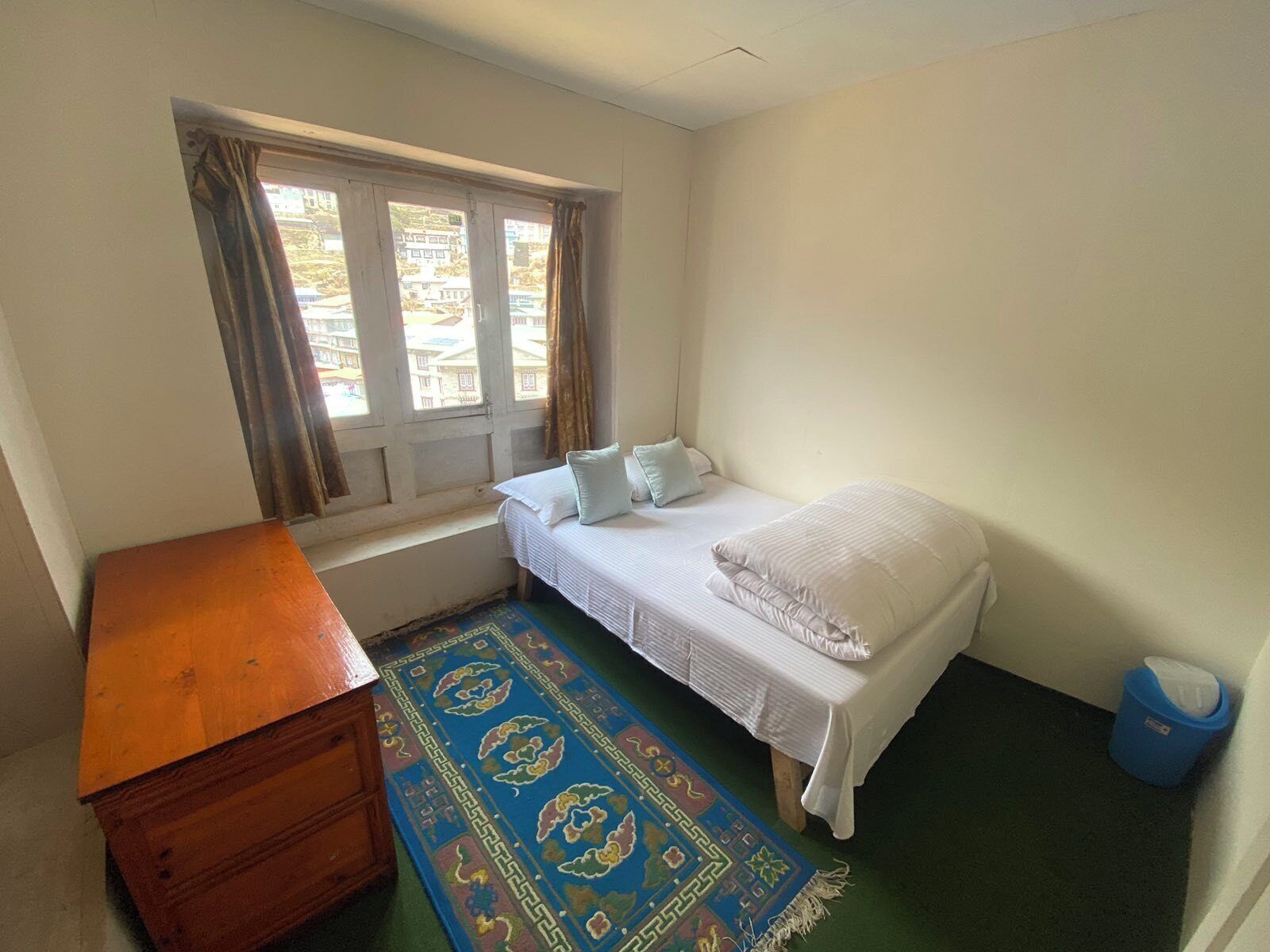 Une petite pièce simple, avec un lit simple, une commode en bois, une fenêtre avec des rideaux et un tapis bleu.