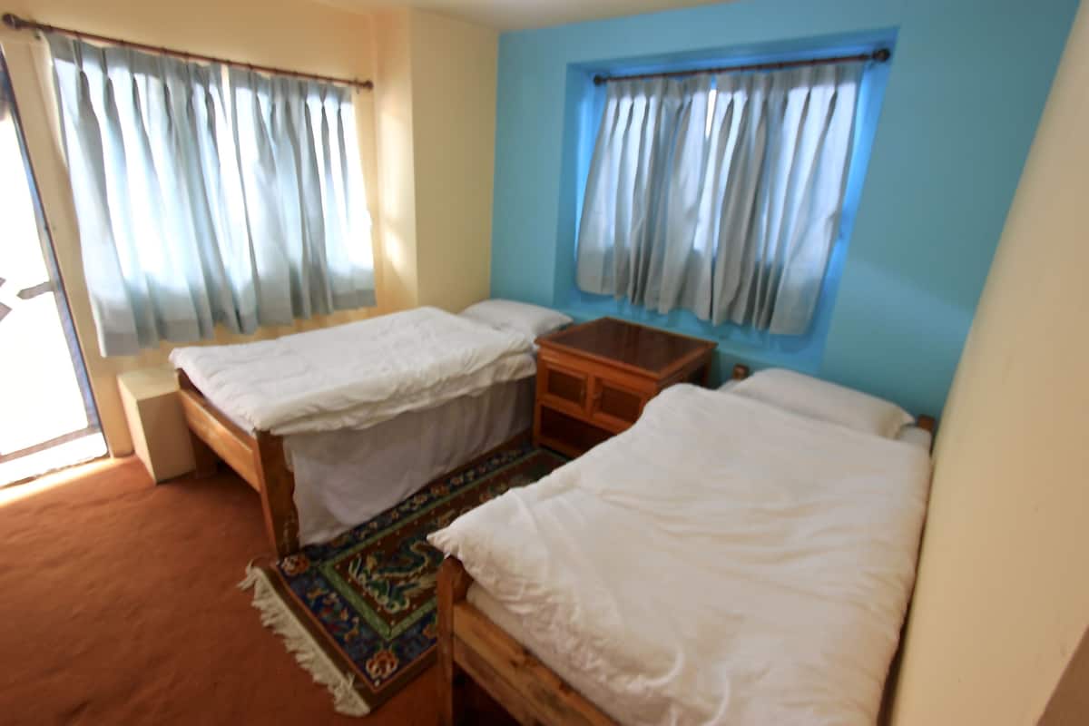 A room with two single beds, a small wooden cabinet, a colorful rug, and a window with curtains.
