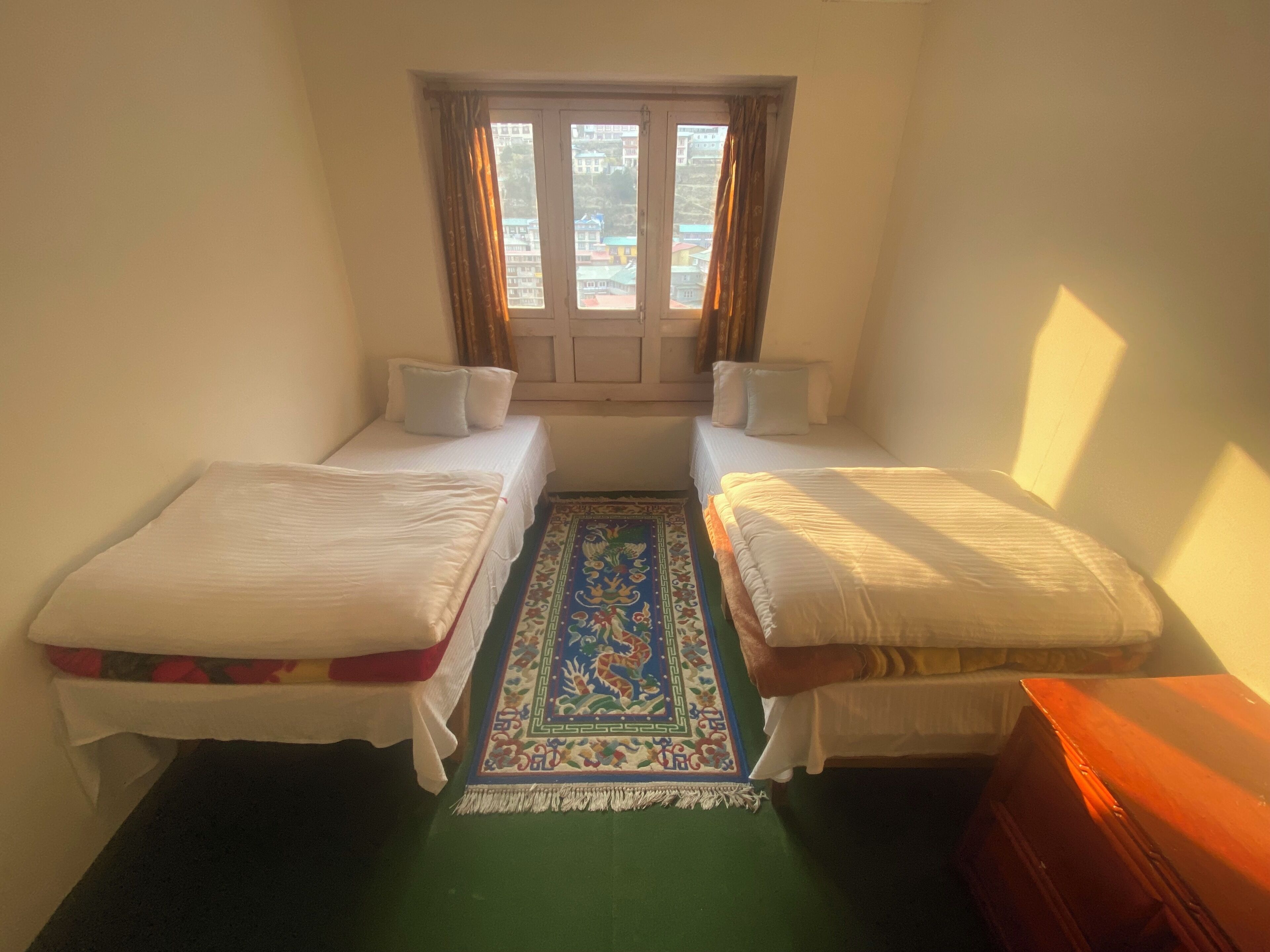 Een kleine kamer met twee eenpersoonsbedden, een raam met gordijnen, een kleurrijk vloerkleed en een houten dressoir.