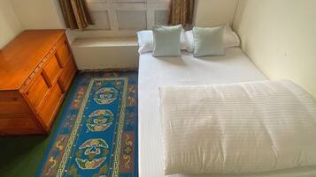 Una habitación con una cama, un cofre de madera, una ventana con cortinas y una alfombra estampada.