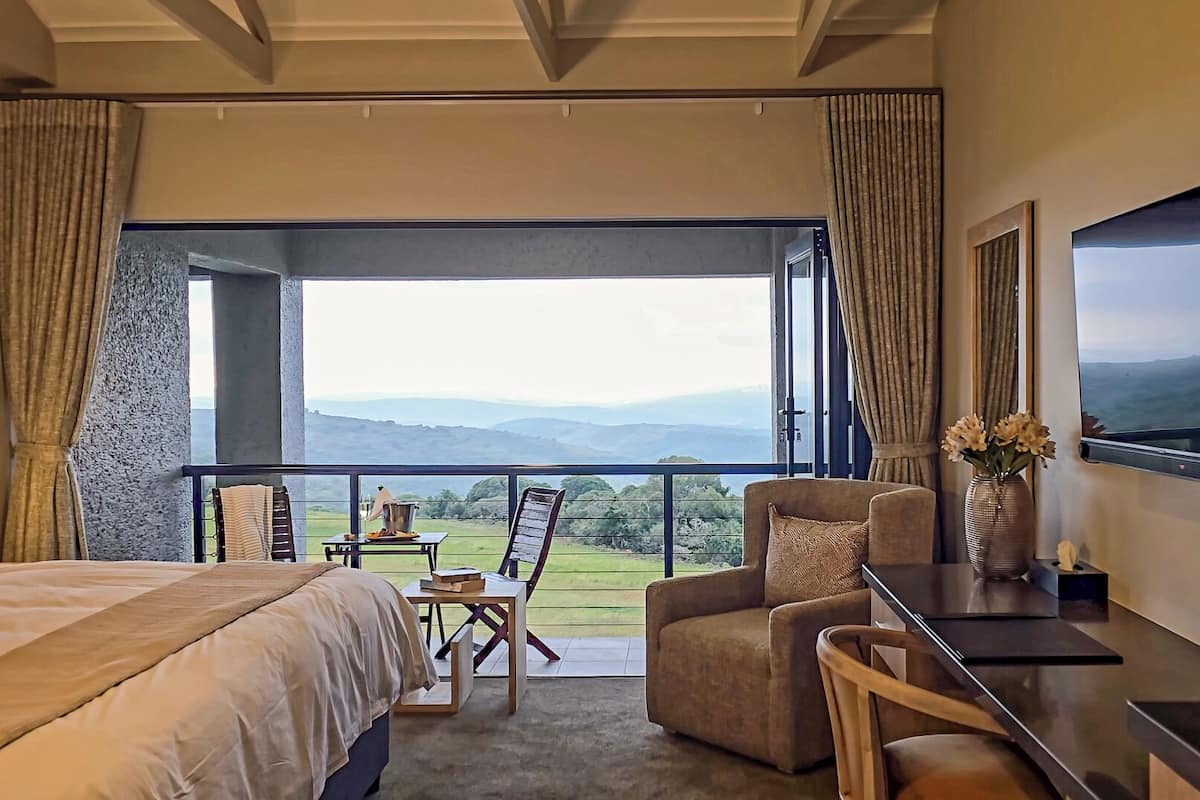 A bedroom with a large bed, a desk, a chair, and a view of a mountainous landscape through a balcony.