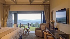 A bedroom with a large bed, a desk, a chair, and a view of a mountainous landscape through a balcony. - Intle Game Lodge (Loerieheuwel)